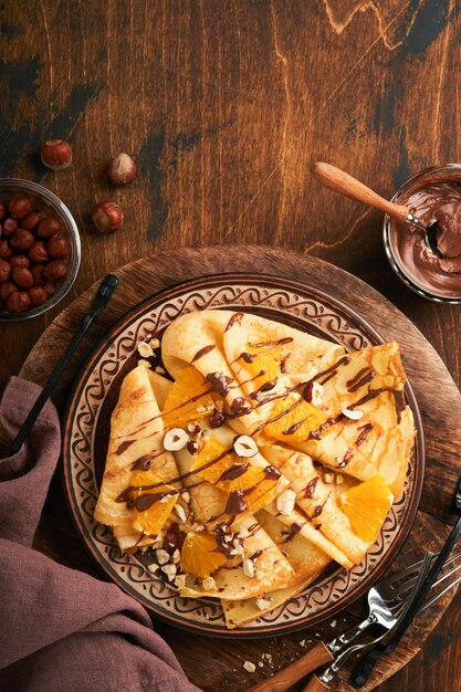 Crepes suzette with oranges and chocolate thin crepes with\
chocolate spread hazelnuts and orange slices fruit in plate for\
breakfast on old dark wooden rustic table background copy space top\
view