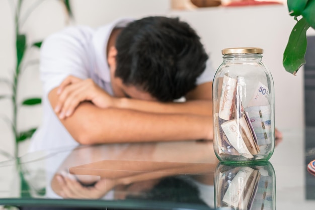 Crestfallen jonge man met zijn spaargeld in een glazen pot op de tafel bankbiljetten in een glazen pot