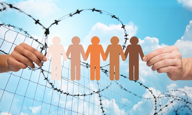 crime, imprisonment, refugee and humanity concept - multiracial couple hands holding chain of paper people pictogram over blue sky and barb wire background