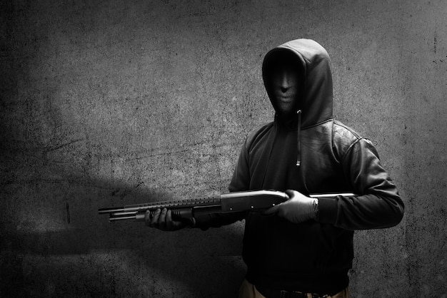 写真 黒い壁の背景と散弾銃を保持している隠されたマスクの犯罪者