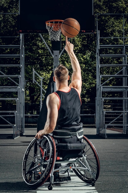 Cripple basketball player in wheelchair plays basketball on open air ground.