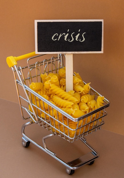 Crisis text on blackboard label against shopping trolley cart\
filled with pasta on beige background food and groceries shopping\
price increase rising food cost food crisis inflation