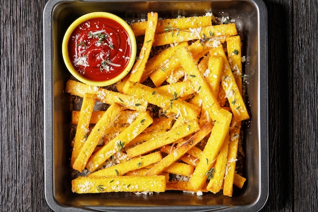 Patatine fritte croccanti di polenta al forno con parmigiano, timo e spezie in una teglia con salsa di pomodoro su una tavola di legno rustica, cucina italiana