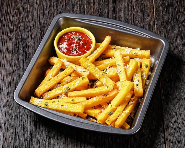 Crispy baked polenta fries with parmesan cheese, thyme and spices in a baking dish with tomato sauce on a rustic wooden table, italy cuisine