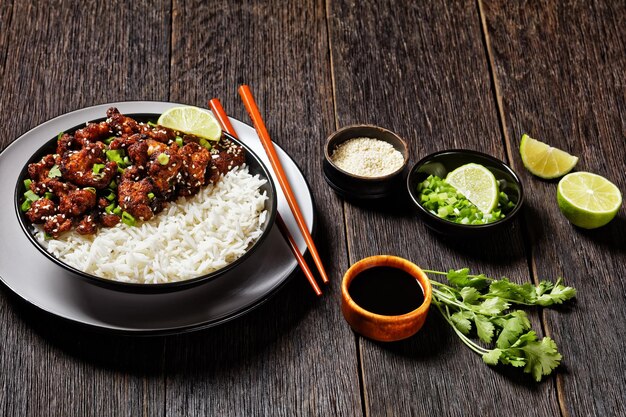 Crispy cauliflower wings with teriyaki sauce topped with fresh\
cilantro, sesame seeds, scallion served with lime and rice on a\
black bowl on a dark wooden table with chopsticks, top view,\
close-up