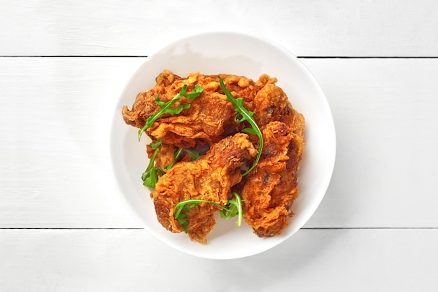 Crispy deepfried chicken wings with fresh arugula in bowl on white wooden table