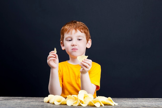 赤い髪の小さな男の子が有害な食べ物を食べるが、子供がテーブルのクローズアップで男の子を本当に食べたいと思っているサクサクのおいしいポテトチップス