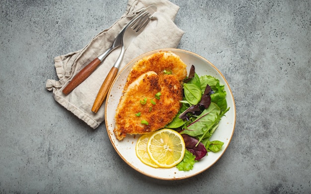 Crispy panko breaded fried chicken fillet with green salad and lemon on plate with cutlery on gray rustic concrete background table from above Japanese style deep fried coated chicken breasts