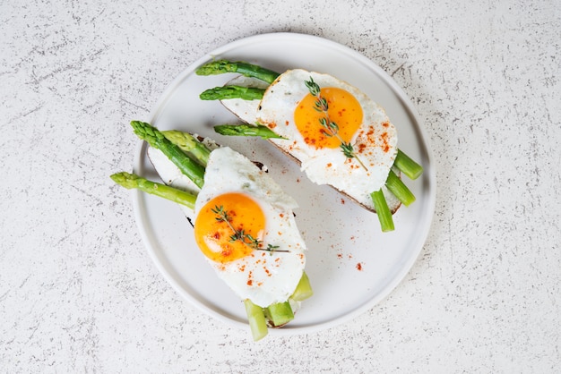 Pane tostato croccante con crema di formaggio cremoso, uovo fritto e asparagi in un piatto su uno sfondo bianco.