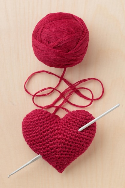 Crocheted amigurumi pink red heart with a crocet hook on a wooden background with a skein of yarn Valentine's day