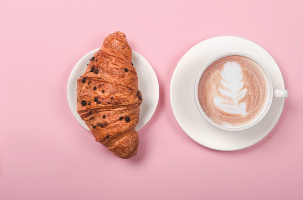 Croissant and cup of coffee on pink background, top view