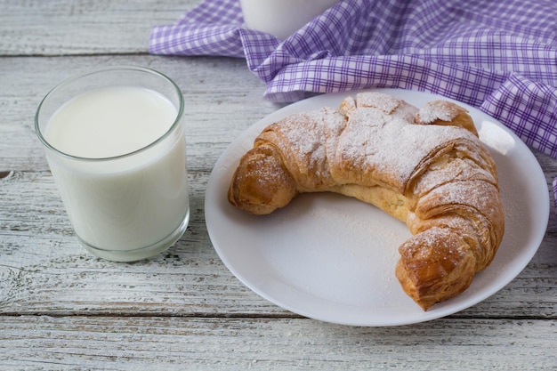 朝食の背景のための古い木製のテーブルの上のミルクと皿の上のクロワッサン