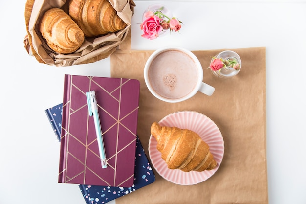 Croissant en een kopje cacao op een witte desktop naast notitieblokken. Break-concept. Bovenaanzicht, plat leggen