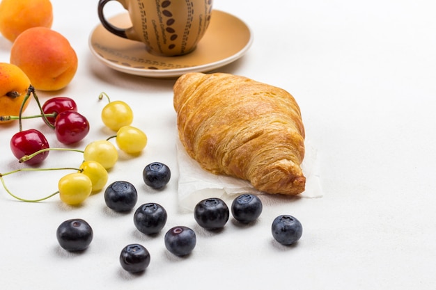 Croissant, kopje koffie, kersen en bosbessen op tafel.