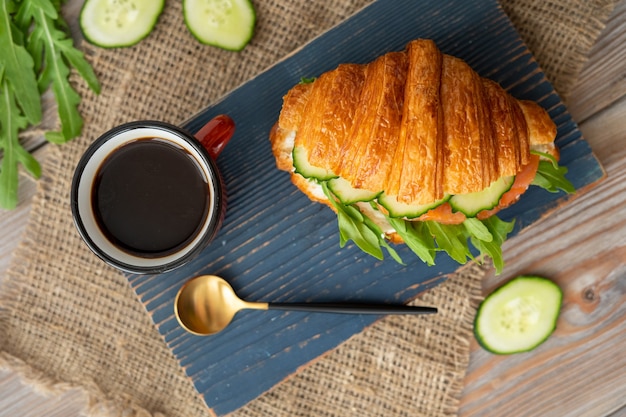 Croissant met komkommer, sla en zalmsandwich met kopje koffie op blauw houten bureau