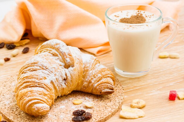 Croissant in powdered sugar and glass of milk wiht heart from cinnamon on a wooden table