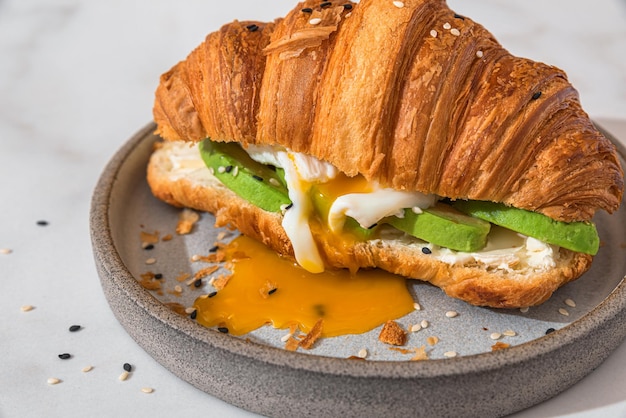 Croissant sandwich with poached egg avocado and soft cheese in a plate on white background French breakfast Tasty food