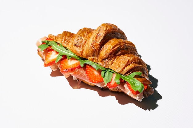 Panino croissant con prosciutto, fragole, rucola e salsa balsamica. luce forte. sfondo bianco