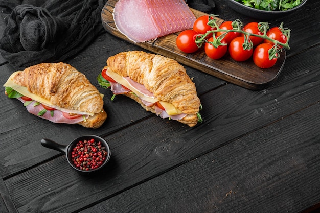 Croissant sandwich with prosciutto, tomatoes, cheese set, with herbs and ingredients, on black wooden table background, with copy space for text