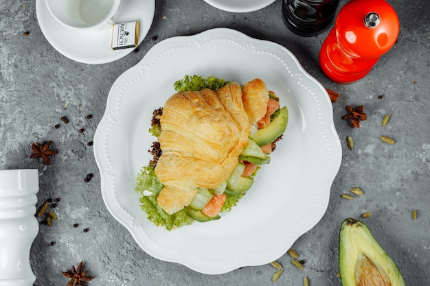 Panino croissant con pesce rosso, avocado, verdure fresche e rucola su tavola di scisto nero su sfondo di pietra nera. concetto di cibo sano.