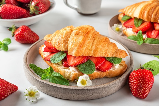 Croissant sandwiches with fresh ripe strawberries and cream cheese in a plate with cup of coffee for tasty breakfast on white table