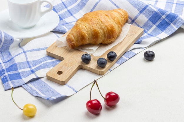 Croissant on white and blue checkered napkin. Cup of coffee cherry and blueberries on table.