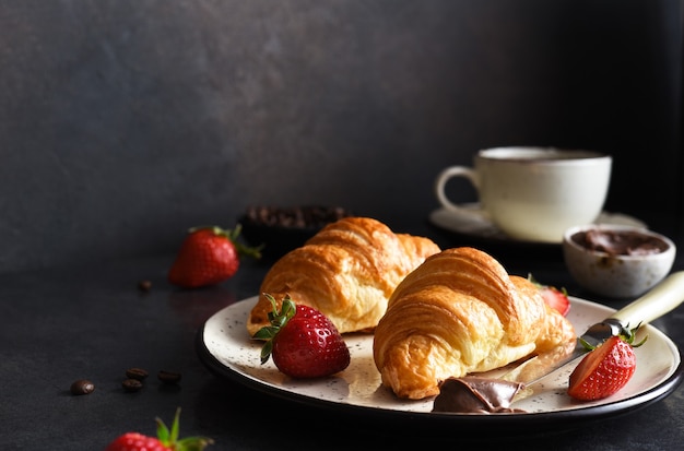 Croissant with chocolate paste and a cup of coffee, strawberries on the kitchen table. âTraditional snack or breakfast.