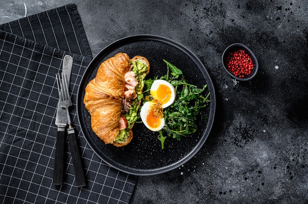 Croissant with hot smoked salmon, avacado, arugula and egg. black background. top view