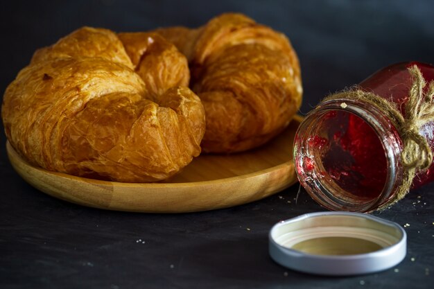 Croissant on wooden dish and strawberry jam bottles in dark background.