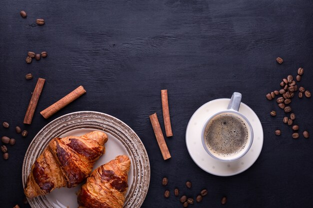 Croissants on a ceramic plate, a cup of coffee, cinnamon sticks and coffee beans on a black wooden table.