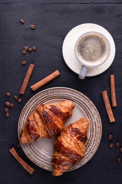 Foto croissants op een keramische plaat, een kopje koffie, kaneelstokjes en koffiebonen op een zwarte houten tafel.