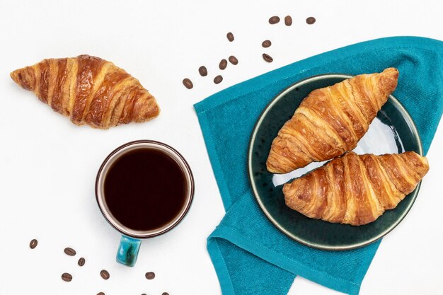 Photo croissants on plate on blue napkin. cup of coffee and grains of coffee on table. flat lay. white background.