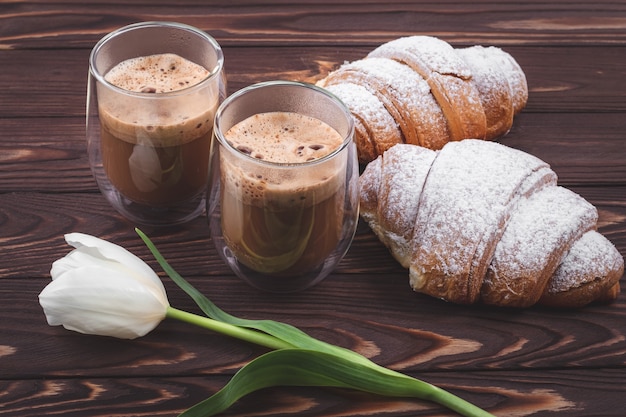 Croissants and two glasses of frothy coffee on a dark brown wooden table. French breakfast concept, break. Drinks and sweets, puff pastry and white flower on boards, spring, morning time.