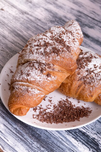 Croissants with powdered sugar and chocolate on white plate on colored wood . Close up.