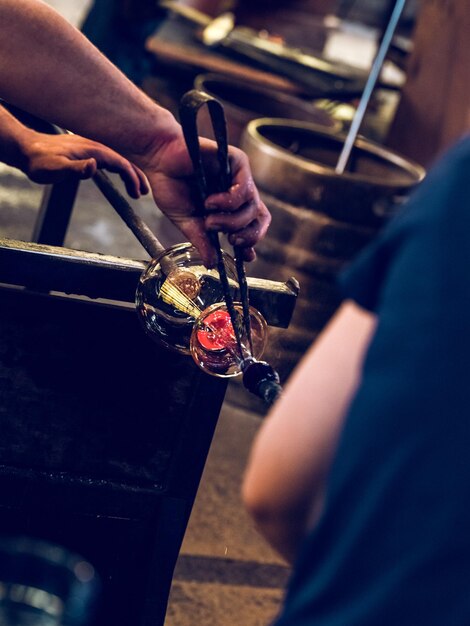 Crop anonymous craftsmen working with hot glass and creating glass bottle fixed on metal bar and using professional tool in glass factory