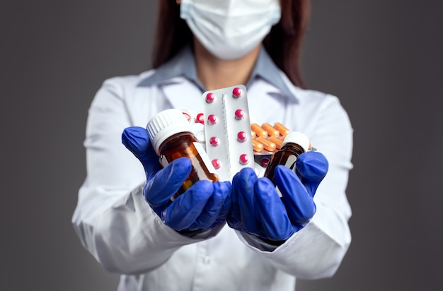 Crop anonymous female medic in protective mask and latex gloves demonstrating heap of various antibiotic pills for viral disease treatment while standing against gray background