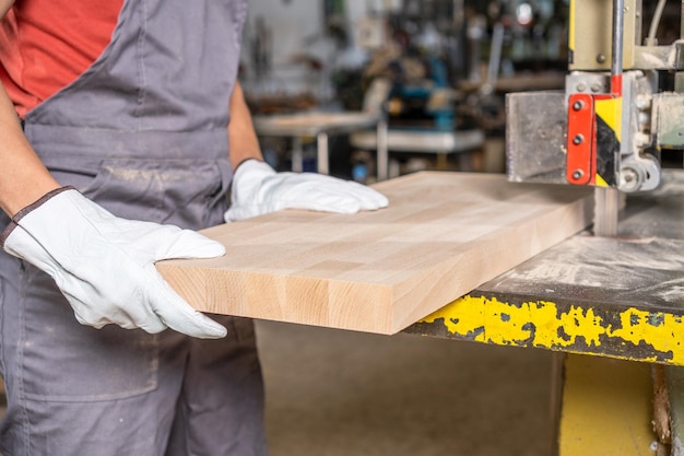 Crop anonymous male artisan in protective wear cutting timber\
board with sharp blade of band saw in professional joinery\
factory