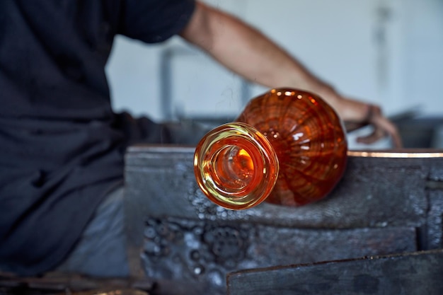 Crop anonymous male forming vase from red hot glass with special instrument in light building with blurred background