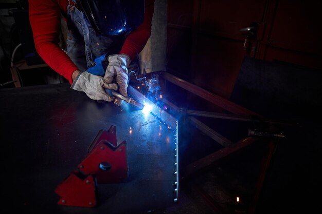 Crop anonymous master in protective helmet welding metal detail to table with special equipment while working in dark professional workshop