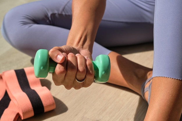 Crop anonymous sportswoman in leggings sitting on floor with small dumbbell in hand during training in sunny day