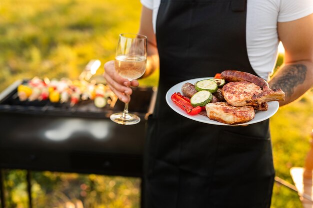 Crop anonymous waiter standing in park with plate of grilled food and wineglass