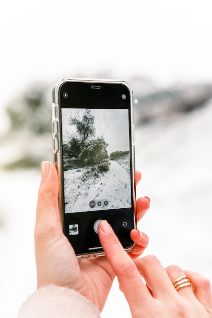 Photo crop anonymous woman taking photo of winter woods covered with snow while using smartphone