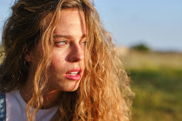 Crop attractive female with long wavy hair and blue eyes spending time on grassy countryside on sunny summer day