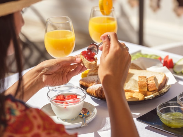 Безобидная женщина-путешественница наносит сладкий варенье на тост, сидя за столом со свежим блюдом и апельсиновым соком на террасе отеля