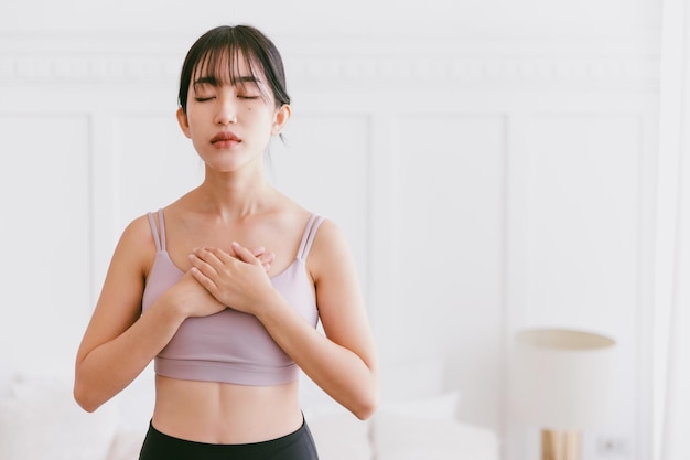スポーティなアジア人女性のクロップイメージの肖像画が胸に手を当て、目を閉じて息を吸い込み、息を吐き出し、リラックスとマインドフルネスを瞑想するヨガの練習をします。