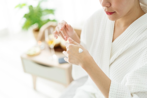 Crop image of a relax young Asian woman in a bathrobe applying moisturizer cream or essential lotion on the back of her hand Woman applying hand cream after bath Seasonal skin protection concept