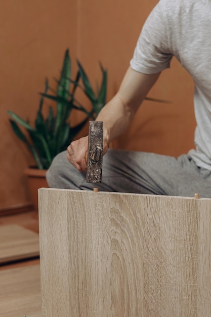 Crop male master hammering wooden dowel into table while assembling new furniture in room at home