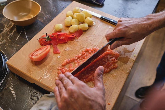 Crop man chopping red tomato carefully on wooden board with lying peeled small potatoes in kitchen