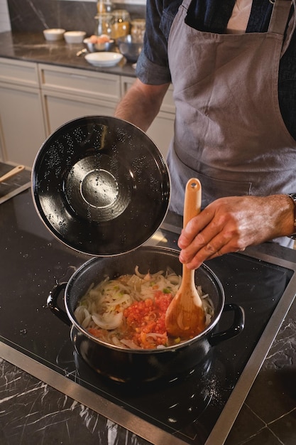 Crop man in gray apron standing at stove and mixing tomato sauce with noodles while cooking in modern kitchen at home