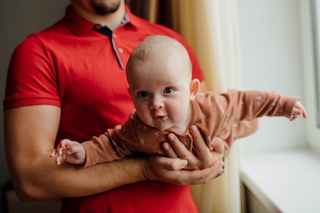 Crop man holding cute curious baby while resting near window in morning at home